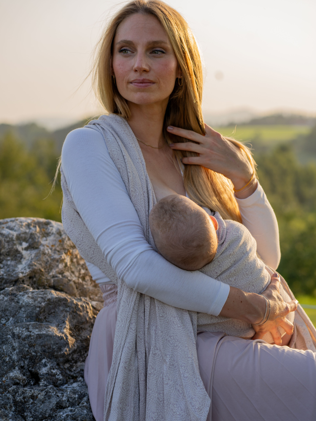 Mutter stillt Baby in Wickelkreuztrage bei Sonnenuntergang auf Felsen