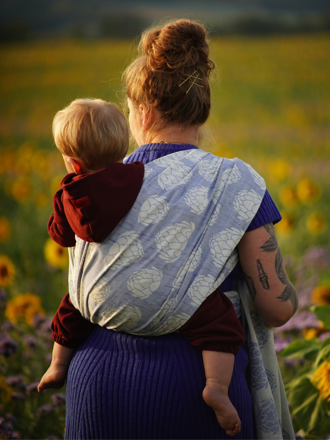 Rückansicht des Artishocca Seren Sky Tragetuchs. Die Mutter steht mitten im Blumenfeld im blauen Strickkleid, das Baby ist auf dem Rücken eingebunden und trägt einen roten Overall